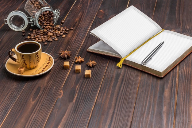 Open notebook with pen Cup of coffee coffee beans in glass jar On the table spices star anise and pieces of brown sugar Dark wooden background Copy space
