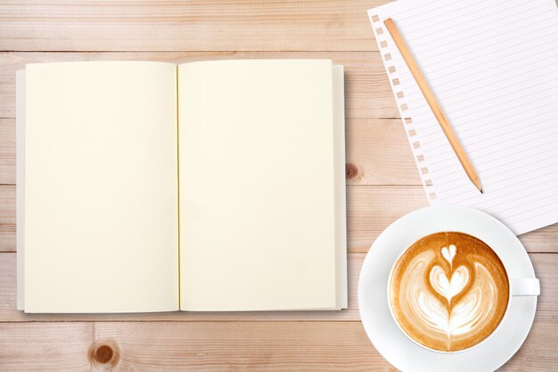 An open notebook with pencil and a cup of coffee on wooden table latte art coffee on top