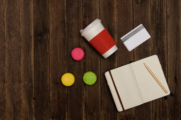 Open notebook with a pencil, macaroons, paper cups and a credit card on a wooden background. Top view, copyspace