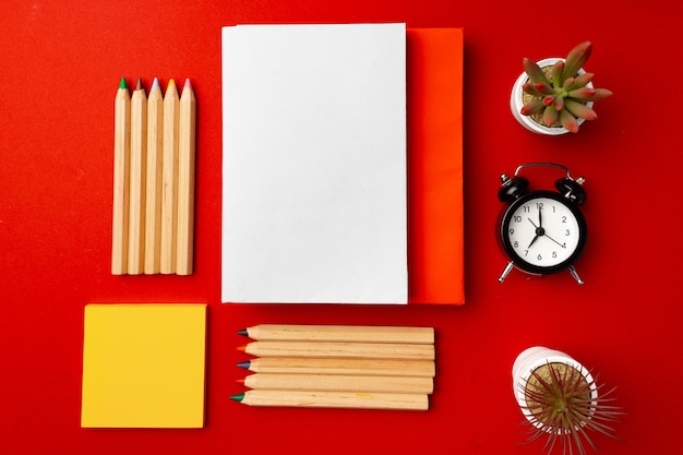 Open notepad with color pencils and flower pots on red background top view