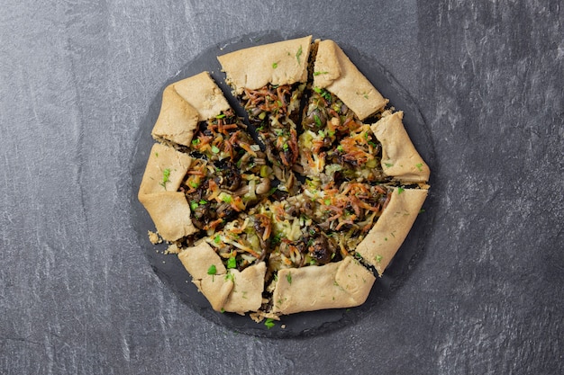 Open pie, galette, with mushrooms, fried green onions, fresh herbs and cheese filling on dark background.