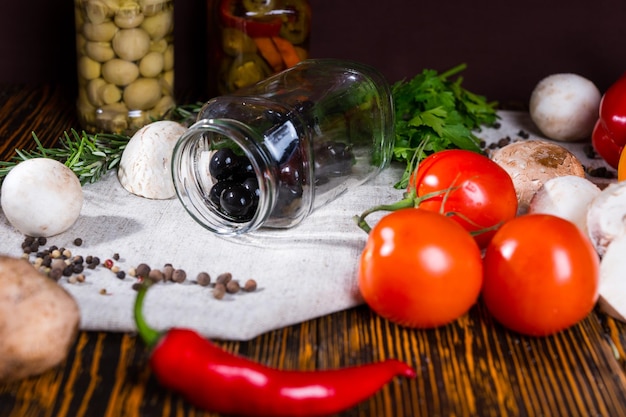 Open pot met zwarte olijven ligt op een servet in de buurt van kruiden, tomaten, champignons en andere groenten op houten tafel