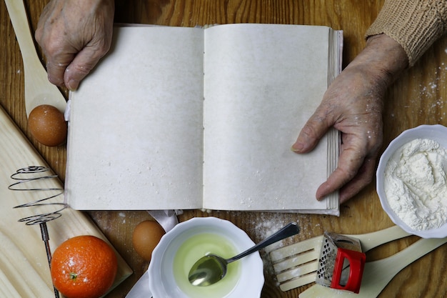 Open recipe book in the hands of an elderly woman in front of a table with utensils