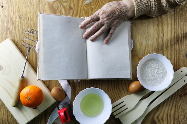 Open recipe book in the hands of an elderly woman in front of a table with utensils