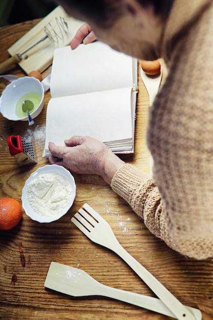 Открытая книга рецептов в руках пожилой женщины перед столом с посудой