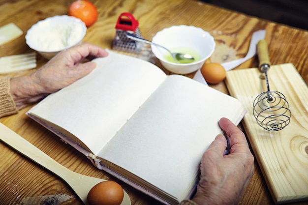 Open recipe book in the hands of an elderly woman in front of a table with utensils