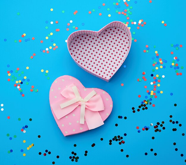 Open roze hartvormige kartonnen doos op een blauwe achtergrond met veelkleurige glanzende confetti