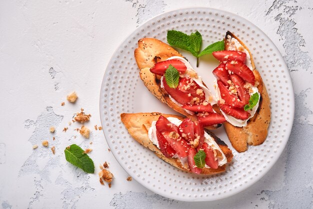 Open sandwiches with strawberries, soft cheese mint and walnut in ceramic plate on a light grey stone background. Summer and healthy dieting food, vegetarian food concept. Top view.