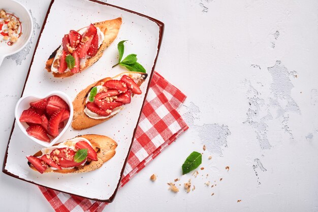 Open sandwiches with strawberries, soft cheese mint and walnut in ceramic plate on a light grey stone background. Summer and healthy dieting food, vegetarian food concept. Top view.