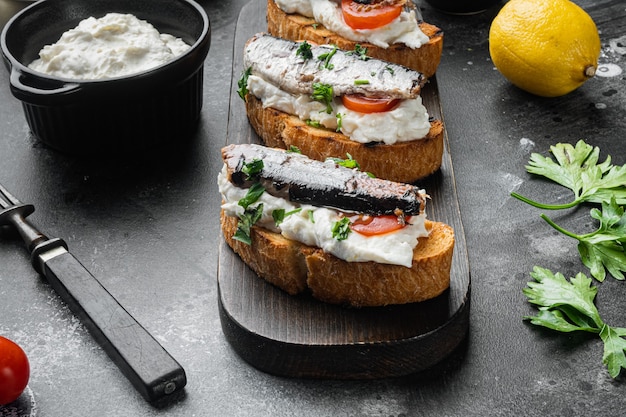 Открытые бутерброды с тостами, рикоттой и рыбными сардинами, на черном фоне темного каменного стола