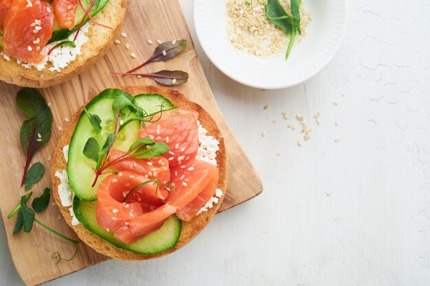 Open smoked salmon sandwiches with cream cheese cucumber sesame seeds microgreens spinach and peas leaves on light old wooden background Healthy breakfast food Delicious snack Top view