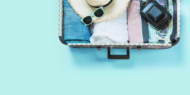 Open suitcase with female clothes for trip on pastel blue. 