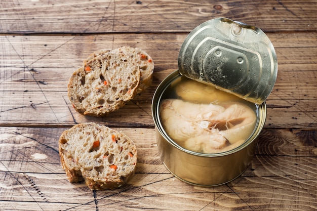 Open tin can with pink salmon fish and pieces of bread on wooden table.