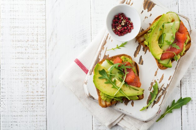 Open toast with trout, salmon, avocado, cucumber and arugula on wooden stand on white wooden table background. Selective focus. Top view. Copy space