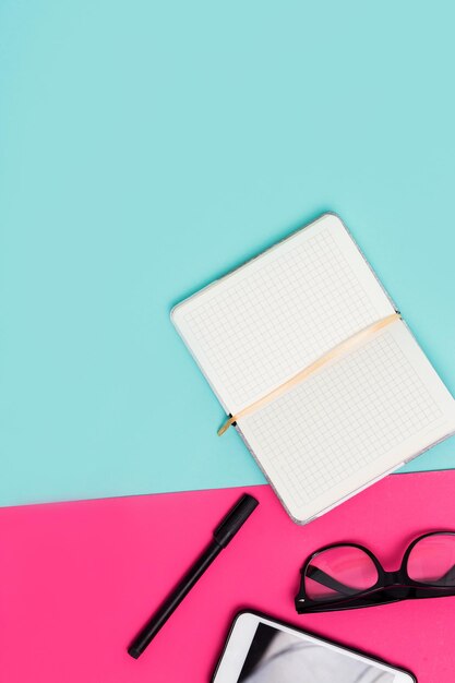 Open white Notepad, glasses, smartphone, pens on colored blue, pink background with copy space. Office items business women