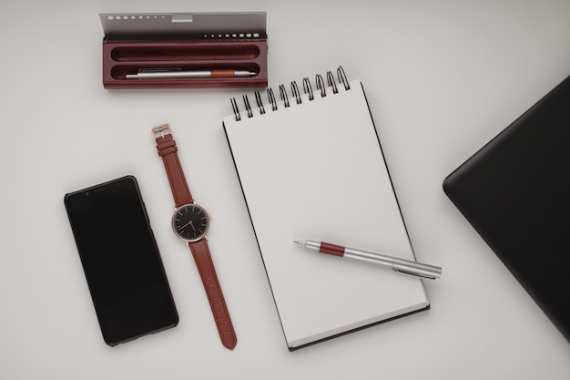 Open white spiral notebook with a blank sheet and a lot of office accessories, a wrist watch, mobile phone , pen case and a laptop.