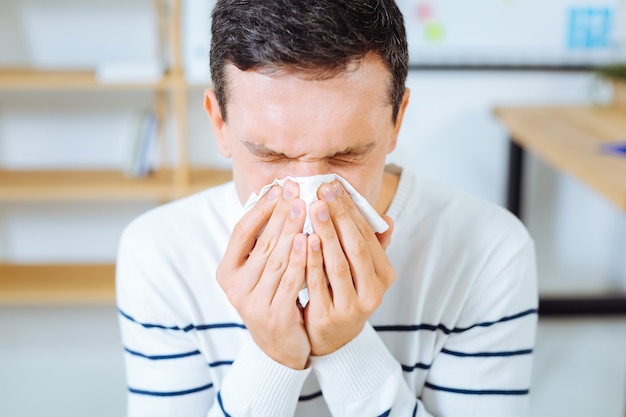 Open your eyes. Indifferent brunette holding napkin in both hands and closing eyes while blowing nose