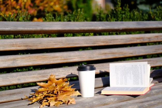 Opened book and cup of coffee on wooden bench in park