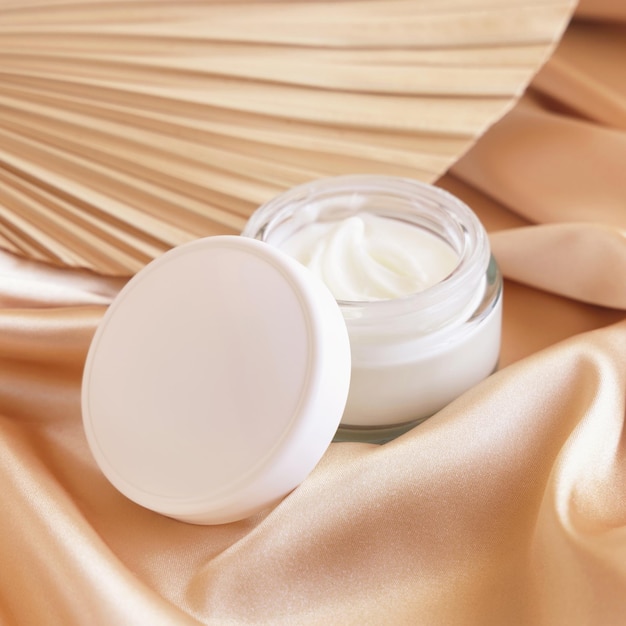 Opened cosmetic jar with white lid on beige satin fabric and dried palm leaf close up mockup