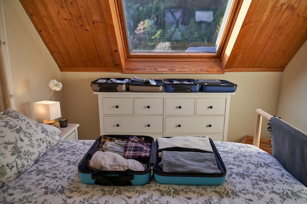 Opened luggage bags with folded clothes placed on comfortable bed with cushion and chest with drawers in modern bedroom