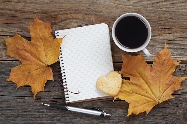 Opened notebook and coffee with cookie in shape of heart and autumn maple leaves on old wooden table, top view