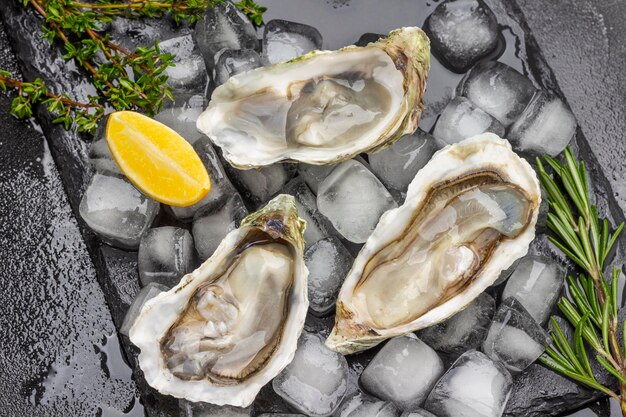 Photo opened oyster shells with ice on black background lemon and greens on table flat lay black background