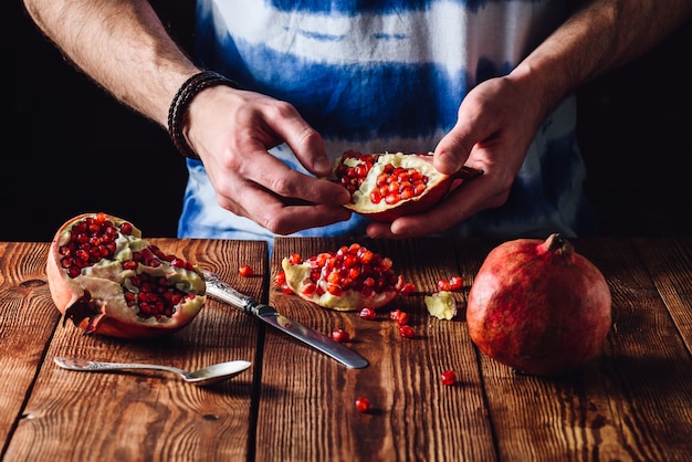 Opened pomegranate fruit in hands and other pieces with knife and spoon on the table