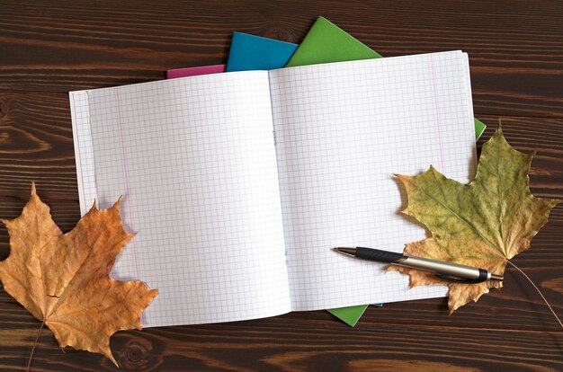 Открытая школьная тетрадь, карандаш и осенние листья на деревянных фоне, вид сверху. Место для текста