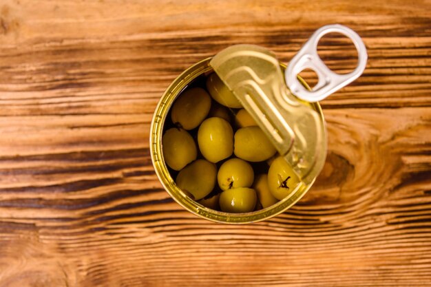 Opened tin can with green olives on wooden table Top view