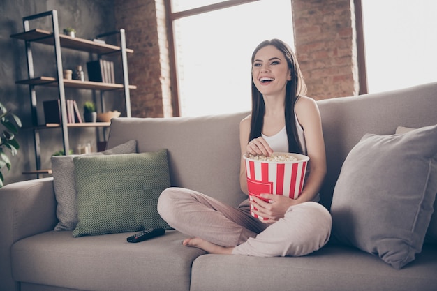 openhartig meisje zit divan benen gekruist kijken naar grappige film eet popcorn