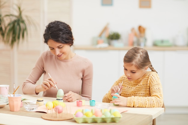 Openhartig portret van moeder en dochter schilderij paaseieren pastelkleuren zittend aan tafel in gezellige keuken interieur, kopie ruimte