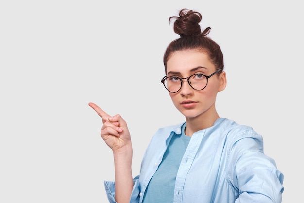 Openhartig schot van serieuze jonge vrouw draagt blauw shirt en ronde transparante bril geeft aan met wijsvinger iets demonstreert op lege kopieerruimte voor advertenties die naar de camera kijken