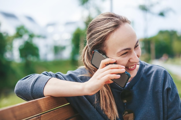 Openhartige jonge vrouw die lacht terwijl ze telefonisch praat, buiten op een bankje zit