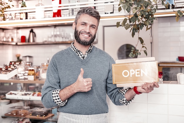 Opening hours. Joyful pleasant man holding a door sign while greeting cafe visitors