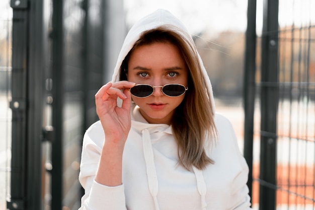 Openlucht dicht omhooggaand portret van jonge mooie vrouw met lang haar in zonnebril, gekleed in een witte hoodiesweater, dichtbij het sportterrein. jeugdcultuur zomer tijdverdrijf