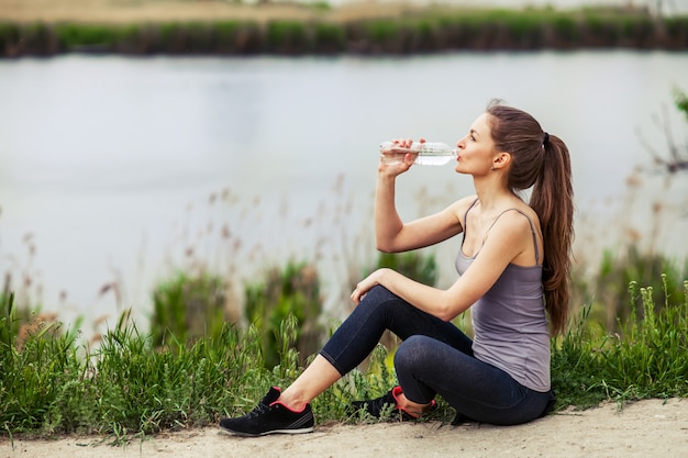 Openluchtfoto van vrij jonge vrouw die sportieve kleren en drinkwaterzitting naast een rivier dragen