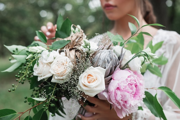 Openluchtportret van de bruid in een witte kanten peignoir houdt in haar handen een huwelijksboeket met rozen