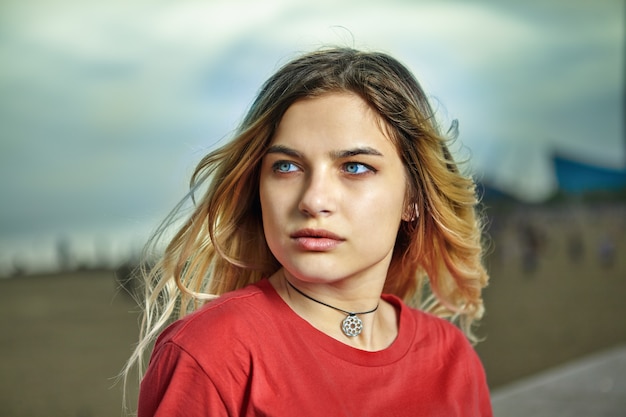 Openluchtportret van ernstig wit meisje ongeveer 21 jaar oud met blond haar en rood overhemd.