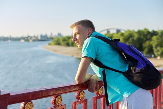 Openluchtportret van knappe tienerjongen 15, 16 jaar oud, met exemplaarruimte. Kerel blond met rugzak kijkt naar de rivier, staande op de brug, zonsondergang zonnige zomerdag, stedelijke levensstijl