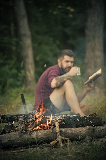 Openluchtrecreatie Hipster-wandelaar met boek en mok bij vreugdevuur in bos