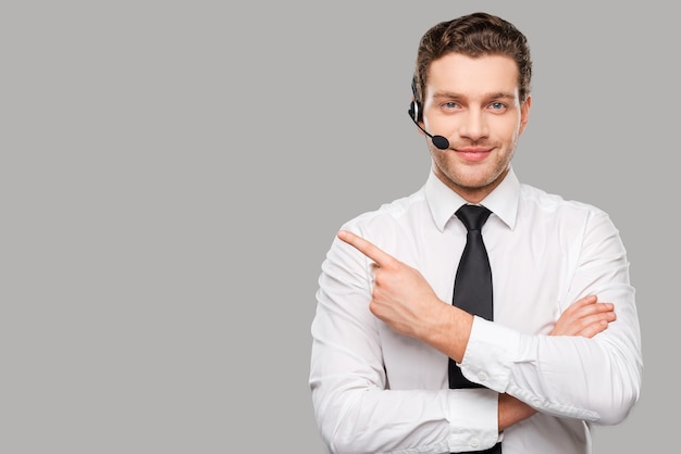 Photo operator pointing copy space. handsome young man in formalwear and headset looking at camera and pointing away while standing against grey background