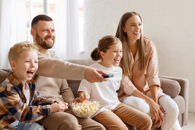 Opgetogen familieouders en kinderen die popcorn eten en lachen terwijl ze op de bank zitten en thuis grappige films kijken