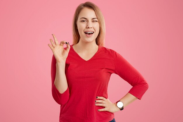 Opgetogen jonge Europese vrouw met vrolijke uitdrukking, toont ok gebaar, knippert oog, houdt hand op taille, gekleed in rode sweater, heeft make-up, geïsoleerd over roze. Ok concept.