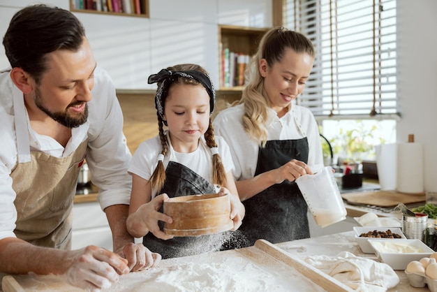 Opgetogen moeder die vloeistoffen in een kom mengt en vader en dochter helpt bij het koken van zelfgemaakte pizza