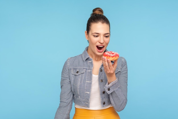 Opgetogen mooie vrouw met haarbroodje die met verlangen naar heerlijke donut kijkt, haar mond openhoudt en droomt om zoete donut te bijten, verleiding van zoete zoetwaren. studio-opname binnenshuis