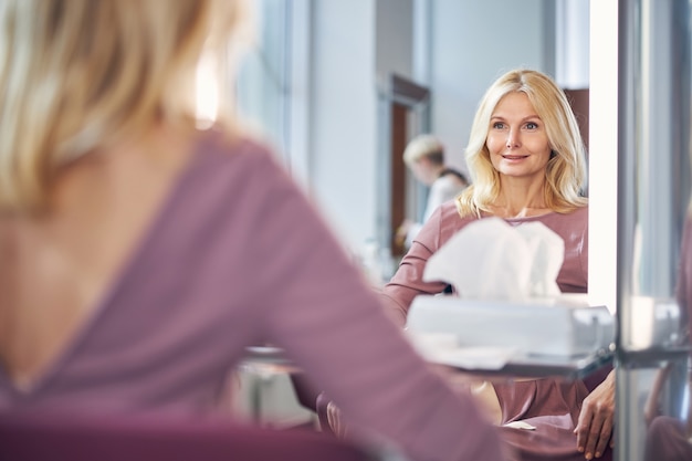 Opgetogen senior vrouw die positiviteit uitdrukt terwijl ze voor een grote spiegel in de schoonheidssalon zit