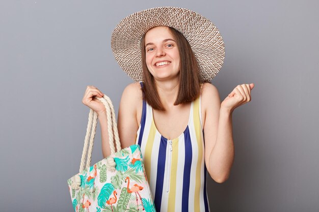 Opgetogen tevreden vrouw met gestreept zwempak en zonnehoed geïsoleerd over grijze achtergrond die zich verheugt over haar zomervakantie die geniet van de zomer