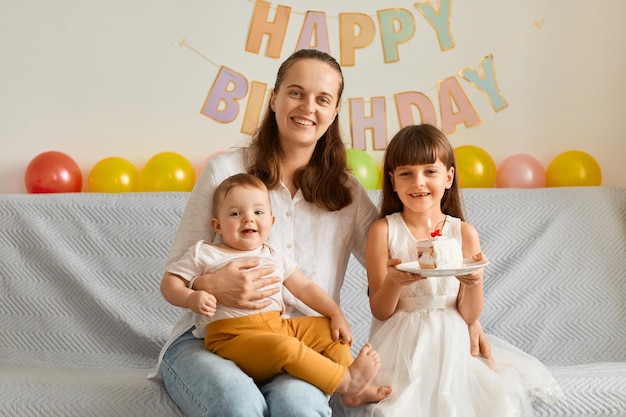 Opgewekte extreem gelukkige kleine meisjes en charmante donkerharige vrouw zittend op de bank met zoete cake glimlachend gelukkig vierend verjaardag samen thuis