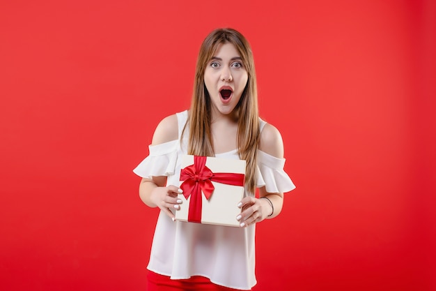 Opgewekte vrouw met giftdoos die witte blouse draagt die op rode muur wordt geïsoleerd