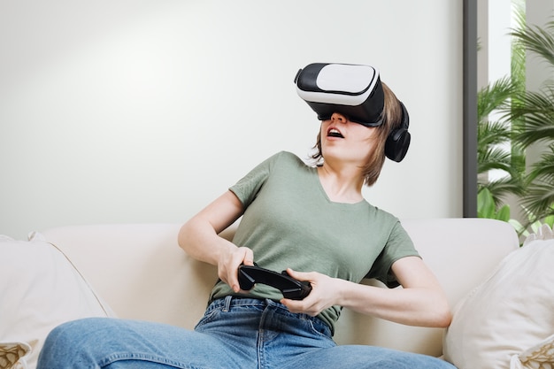 Opgewonden aantrekkelijke vrouw gamen met virtual reality-headset of bril en zittend op de bank, gamepad-controller vasthoudend en videogame spelend. Virtual reality-concept, vrouwen in gaming-concept